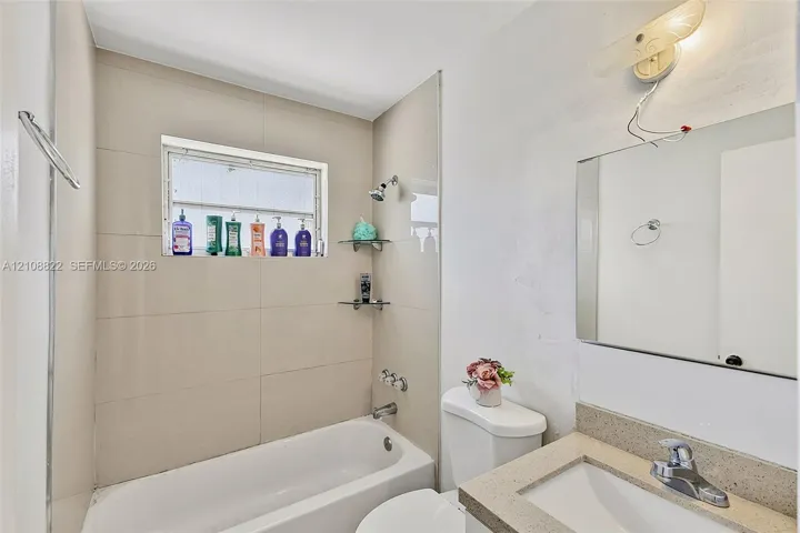 Full bathroom featuring large-format wall tile, a built-in bathtub with showerhead, integrated glass corner shelving, a vanity with a solid surface countertop, and a wall-mounted light fixture