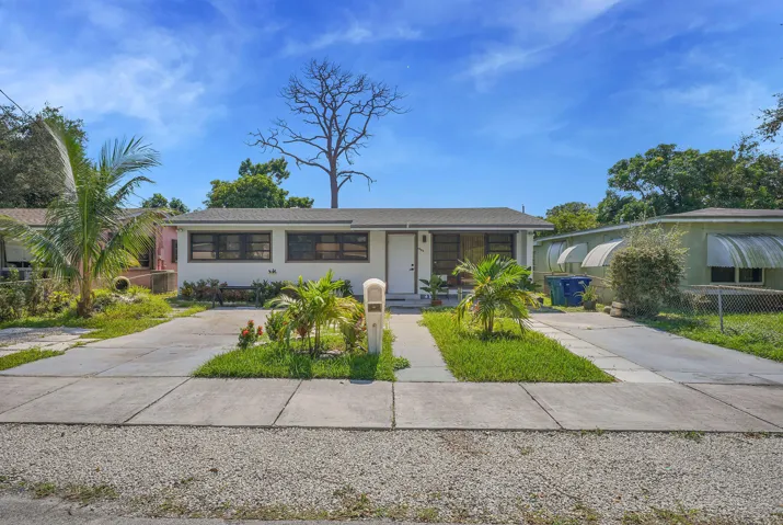 Single-story exterior featuring a light-colored facade, dual concrete driveways, and established tropical landscaping