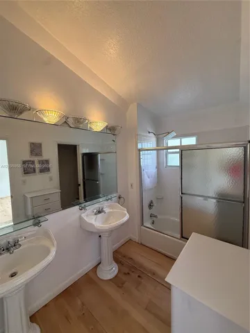 Dual porcelain pedestal sinks, wood-finish flooring, full-width wall mirror, and a tub-shower combination with sliding glass doors