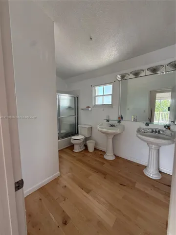 Bathroom featuring wood-finish flooring, dual pedestal sinks, a large wall-mounted mirror with integrated lighting, and a glass-enclosed shower