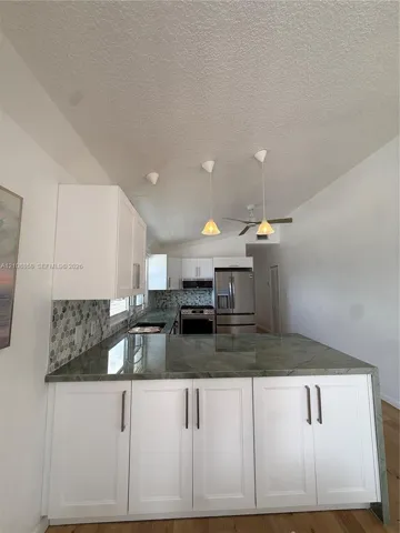 Kitchen featuring white shaker-style cabinetry, solid-surface countertops with waterfall edge, geometric tile backsplash, stainless steel appliances, and pendant lighting