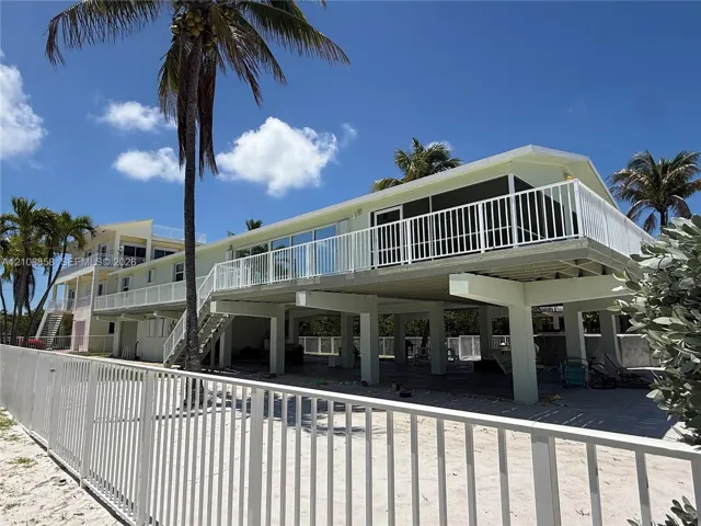 Elevated exterior featuring white metal railings, a screened balcony, concrete pilings, and a sandy ground-level area