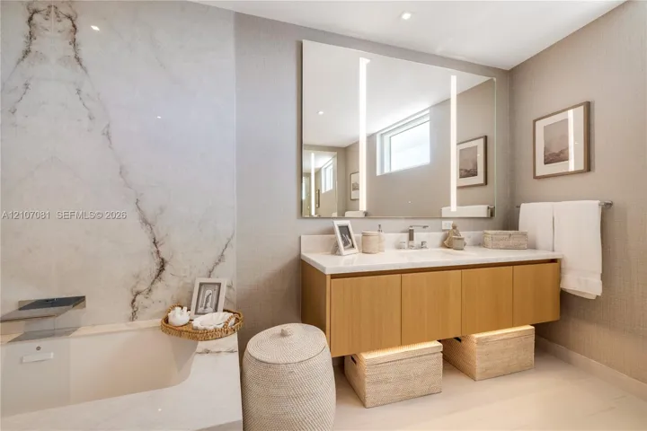 Floating wood-finish vanity with a stone countertop, wall-mounted faucet, integrated mirror with vertical LED lighting, and full-height stone tile surround at the soaking tub