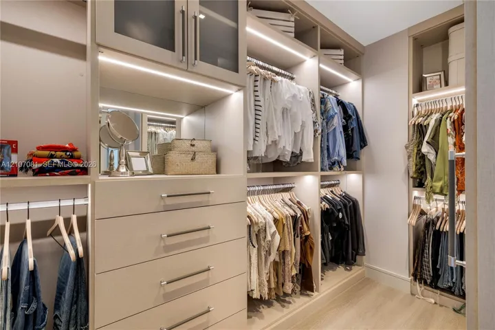 Custom-built walk-in closet featuring floor-to-ceiling cabinetry, integrated LED lighting, glass-front upper storage, soft-close drawers with metallic pulls, and wood-finish flooring