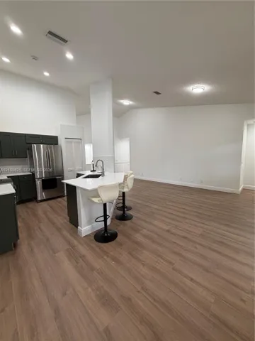 Kitchen featuring wood-finish flooring, dark cabinetry, stainless steel refrigerator, white countertops, and a central island with a built-in sink and faucet