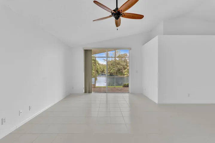 Vaulted ceiling featuring a central five-blade wood-finish ceiling fan, white tile flooring, and glass sliding doors revealing a screened-in lanai with water and natural scenery