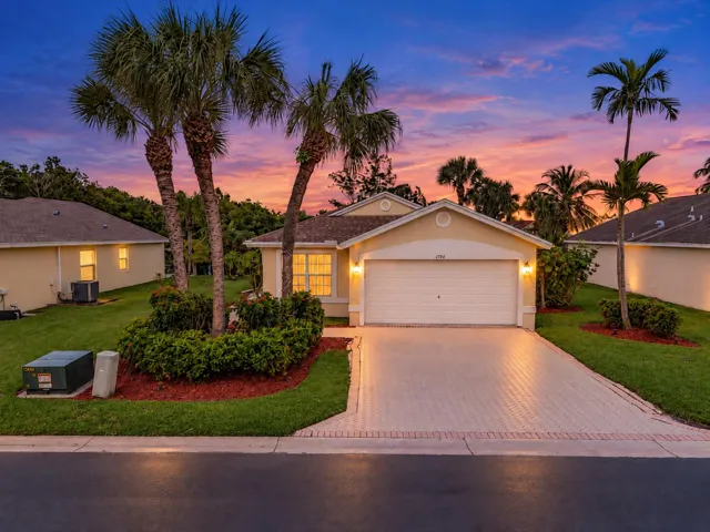Twilight photo - Single-story residence featuring a paved driveway, two-car garage, manicured lawn, and mature palm landscaping