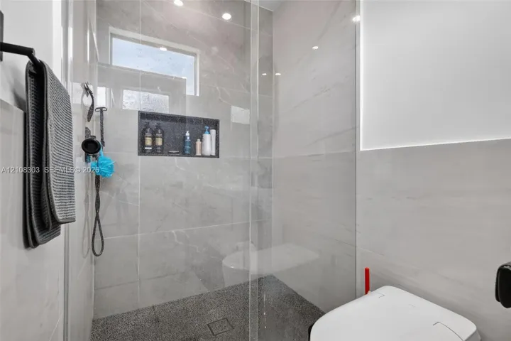 Walk-in shower featuring large-format stone-finish tile surround, integrated recessed storage niche, pebble-textured flooring, frameless glass enclosure, and transom window