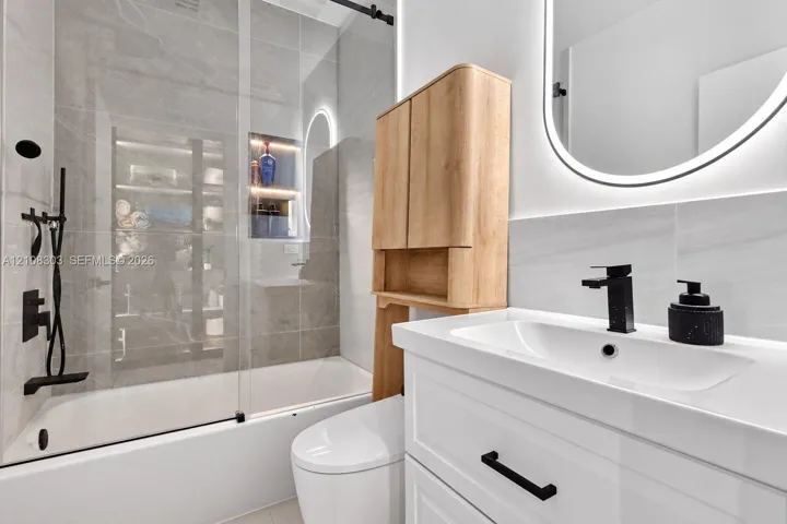 Bathroom featuring a glass-enclosed shower with large-format tile surround, recessed niche lighting, white vanity cabinet with matte black hardware, integrated vanity mirror with LED illumination, and wood-finish cabinetry