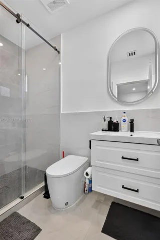 Walk-in shower with a sliding glass door, white wall-mounted vanity with two drawers, matte black hardware, oval-framed mirror, and large-format tile flooring