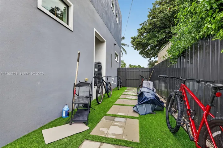 Side exterior featuring grey stucco siding, synthetic turf ground cover, concrete paver pathway, and vertical metal privacy fencing