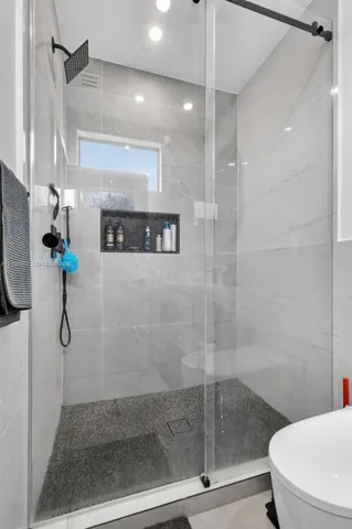 Walk-in shower featuring large-format light grey wall tile, a textured pebble-style floor, a built-in recessed niche, a frameless glass enclosure, and a matte black rainfall showerhead