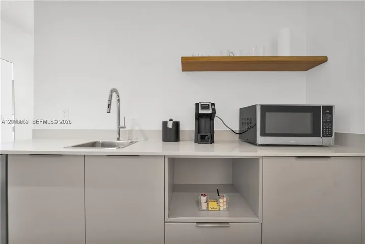 Modern kitchenette featuring flat-panel cabinetry, light-toned countertop, stainless steel undermount sink with gooseneck faucet, and floating wood-finish shelving
