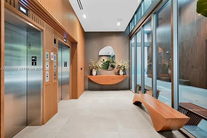 Lobby featuring dual stainless steel elevators, floor-to-ceiling glass paneling, wood-slat wall paneling, built-in curved wood console, and large-format porcelain tile flooring
