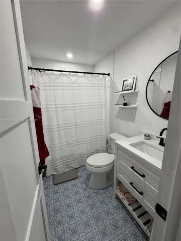 Bathroom featuring patterned tile flooring, white vanity with integrated sink and matte black hardware, round vanity mirror, recessed ceiling lighting, and wall-mounted shelving