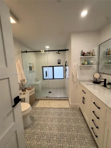 Custom cabinetry with dark hardware, stone-top vanity, walk-in glass-enclosed shower with matte black fixtures, patterned tile flooring, and integrated wall shelving