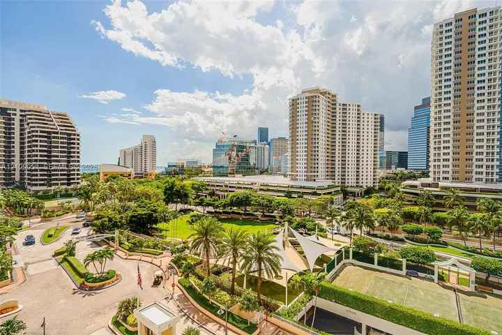 High-rise urban landscape featuring manicured lawns, mature palm trees, tensile shade structures, and an on-site tennis court