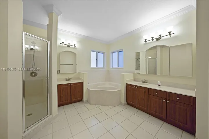 Bathroom featuring dual wood-finish vanities with solid-surface countertops, a corner soaking tub, a walk-in shower with glass enclosure, and ceramic tile flooring