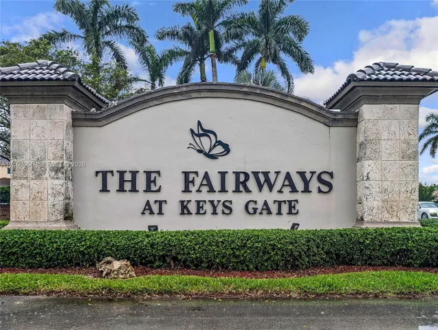 Monument-style neighborhood signage featuring stone-clad columns, arched stucco wall, and mature palm tree landscaping