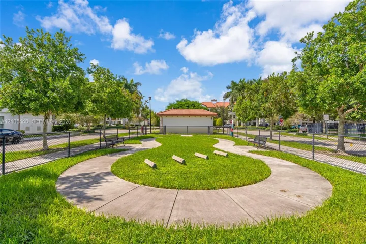 Fenced outdoor recreation area featuring a concrete walking path, manicured lawn, perimeter benches, and mature trees