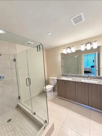 Bathroom featuring a dual-sink vanity with stone-finish countertops, frameless glass shower enclosure, large wall-mounted vanity mirror, and neutral tile flooring