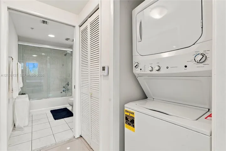 Stacked laundry unit with white finish, closet with louvered doors, bathroom featuring a tub-shower combination with a glass enclosure, and ceramic tile flooring