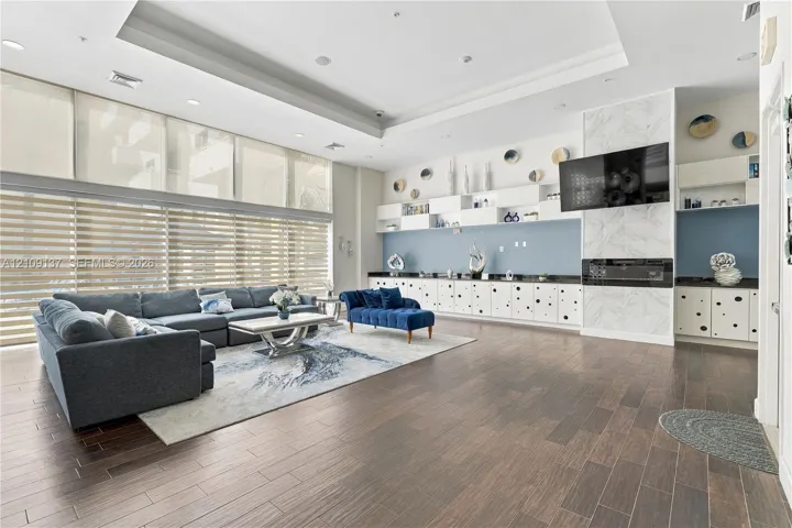 Expansive living space featuring wood-finish flooring, floor-to-ceiling windows with roller shades, recessed tray ceilings, built-in cabinetry, and a fireplace with stone-finish surround