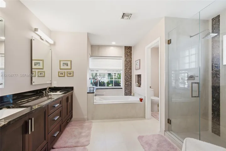 Double vanity with dark wood cabinetry and stone countertops, oversized walk-in glass shower with tile inlay, and built-in soaking tub