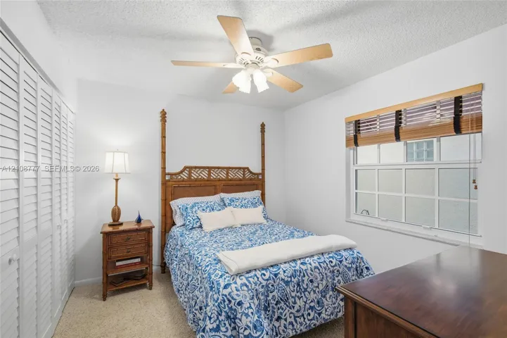Bedroom featuring wall-to-wall louvered closet doors, multi-pane window with wood-slat blinds, textured ceiling, and a central ceiling fan with integrated light fixture