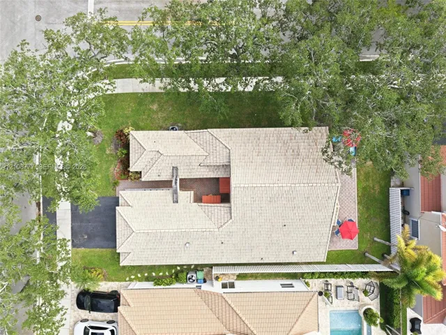 Aerial perspective of a property featuring a tile roof, paved driveway, brick-paver patio, and mature trees on a corner lot