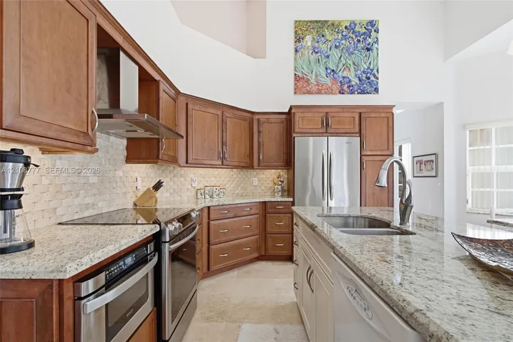 Kitchen featuring custom wood cabinetry, natural stone countertops, a multi-textured stone backsplash, stainless steel appliances, and high ceilings