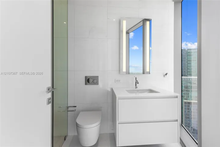 Modern wall-mounted floating vanity with integrated rectangular sink and stone countertop, illuminated rectangular mirror, floor-to-ceiling windows, and wall-mounted dual-flush toilet