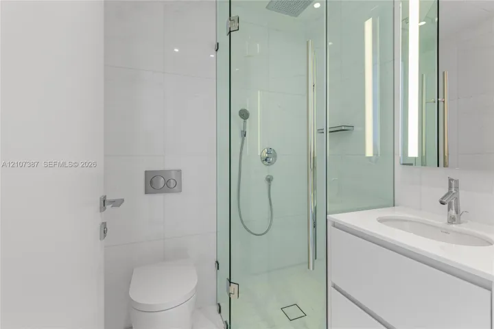 Bathroom featuring a glass-enclosed shower with a rainfall showerhead, wall-mounted dual-flush toilet, white vanity with an integrated sink, polished chrome fixtures, and large-format tile surround