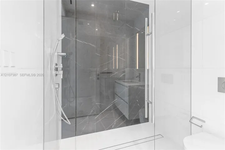 Walk-in shower featuring large-format dark veined tile surround, frameless glass enclosure, handheld shower fixture, and linear drain