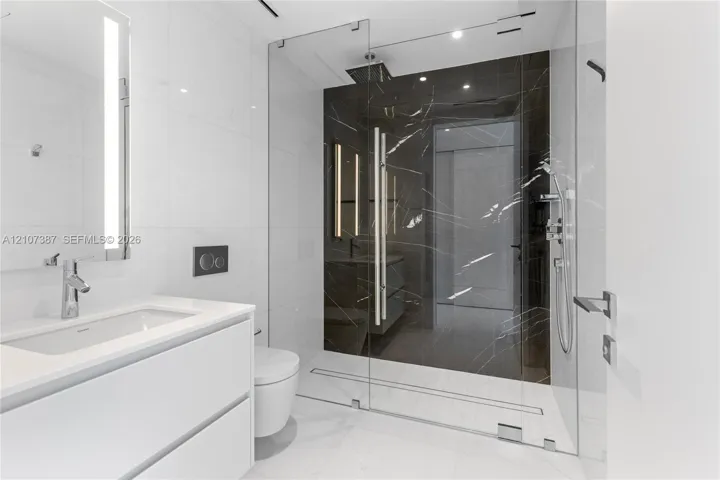 Modern bathroom featuring a floating white vanity with a rectangular sink, wall-mounted faucet, walk-in shower with floor-to-ceiling glass enclosure, dark stone-finish shower surround, and integrated lighting