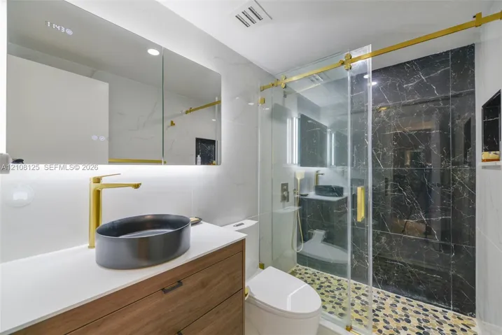 Bathroom featuring a wood-finish vanity with a quartz countertop, oval vessel sink, gold-toned hardware, and a walk-in shower with stone-tile flooring and dark accent tiling