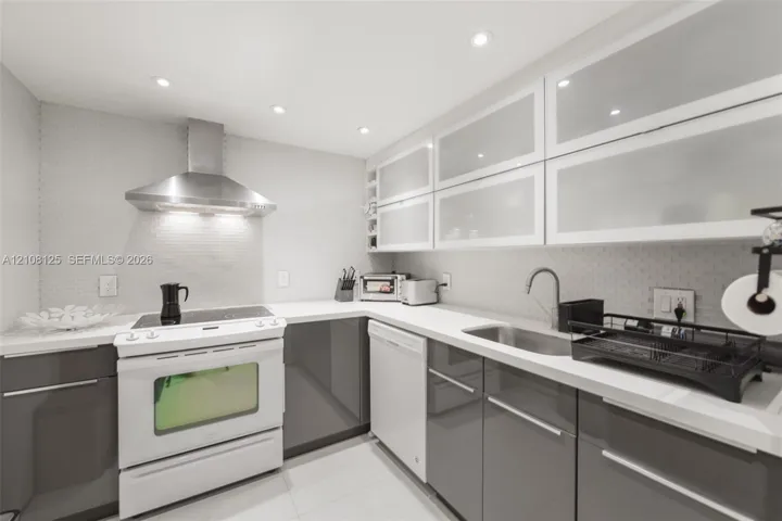 Kitchen featuring high-gloss gray lower cabinetry, white upper cabinetry with frosted glass inserts, stone countertops, stainless steel range hood, and integrated recessed lighting