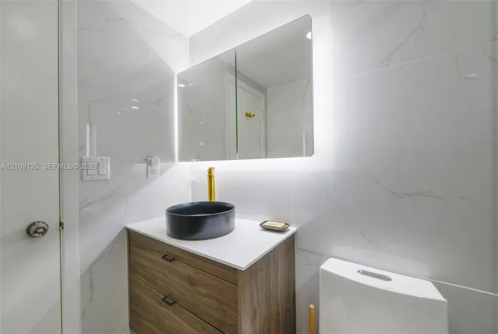 Modern vanity with wood-finish cabinetry, white countertop, black vessel sink, and brass faucet, complemented by wall-to-wall light-toned tiling and an integrated backlit medicine cabinet