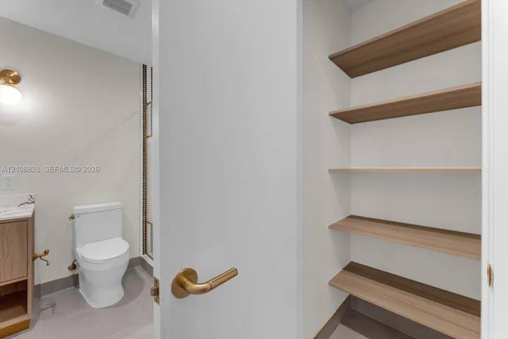 Built-in storage closet featuring tiered wood-finish shelving, brass hardware, neutral-toned cabinetry, and minimalist wall-mounted lighting