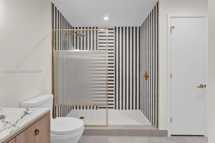 Walk-in shower featuring geometric black-and-white tile, brushed gold-finish hardware, glass panel enclosure, and stone-patterned vanity countertop