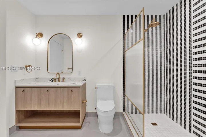 Bathroom featuring a wood-finish vanity with a stone-top counter, arched mirror, brass-toned fixtures, and a shower with a floor-to-ceiling geometric tile surround and textured glass enclosure