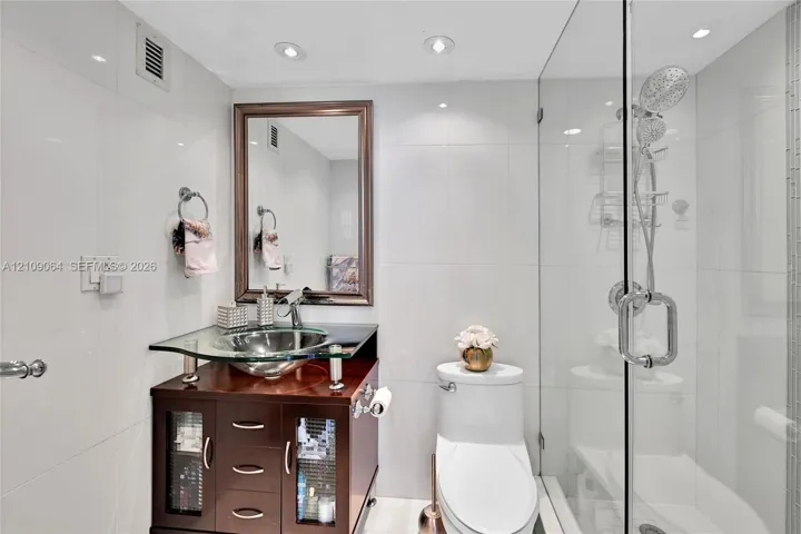 Dark wood-finish vanity with a glass countertop, integrated vessel sink, frameless glass shower enclosure, and recessed lighting