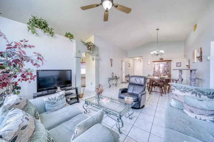Living area featuring vaulted ceilings, tile flooring, an arched architectural wall pass-through, a ceiling fan with light fixture, and a multi-arm chandelier