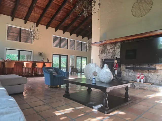 Vaulted ceiling with exposed wood beams, floor-to-ceiling stone fireplace, built-in wooden bar, and tile flooring