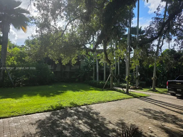 Expansive grounds featuring a paved brick driveway, mature canopy trees, manicured lawn, and established tropical landscaping