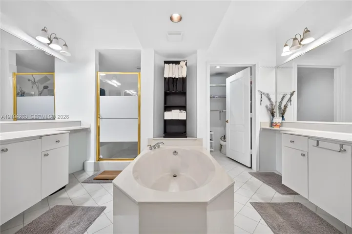 Bathroom featuring dual white vanities with solid-surface countertops, a center soaking tub, a glass-enclosed shower with metallic framing, and tile flooring