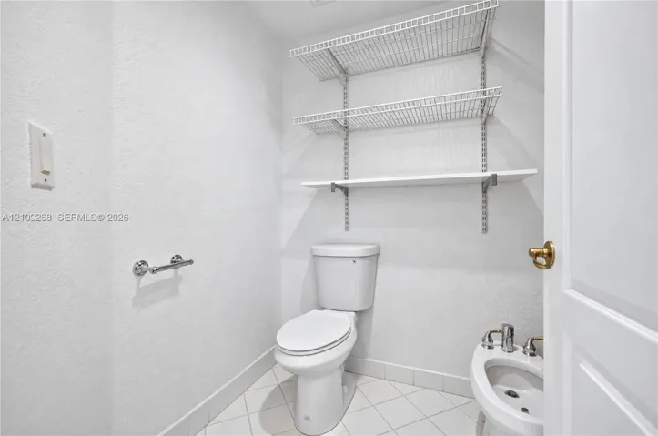 White porcelain toilet, bidet with metal hardware, tiled flooring, and adjustable wall-mounted wire shelving