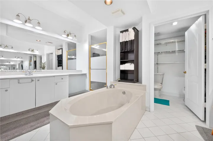 Large soaking tub with a solid surface surround, expansive wall-to-wall mirror, dual-light vanity fixtures, and white tile flooring