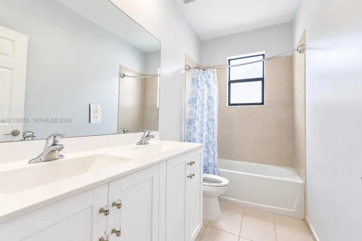 Bathroom featuring a white double-sink vanity with clear acrylic and metal hardware, a wall-to-wall mirror, ceramic tile flooring, and a tub-shower combination with neutral tile surround