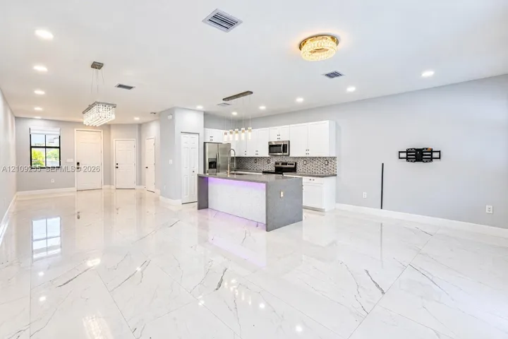 Open-concept kitchen and living area featuring large-format veined tile flooring, a center island with integrated lighting, white shaker-style cabinetry, a geometric tile backsplash, and tiered pendant lighting